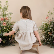 Load image into Gallery viewer, a young girl in natural colour 100% linen day dress with four buttons down back with puff sleeve. Flowers with beige background. 
