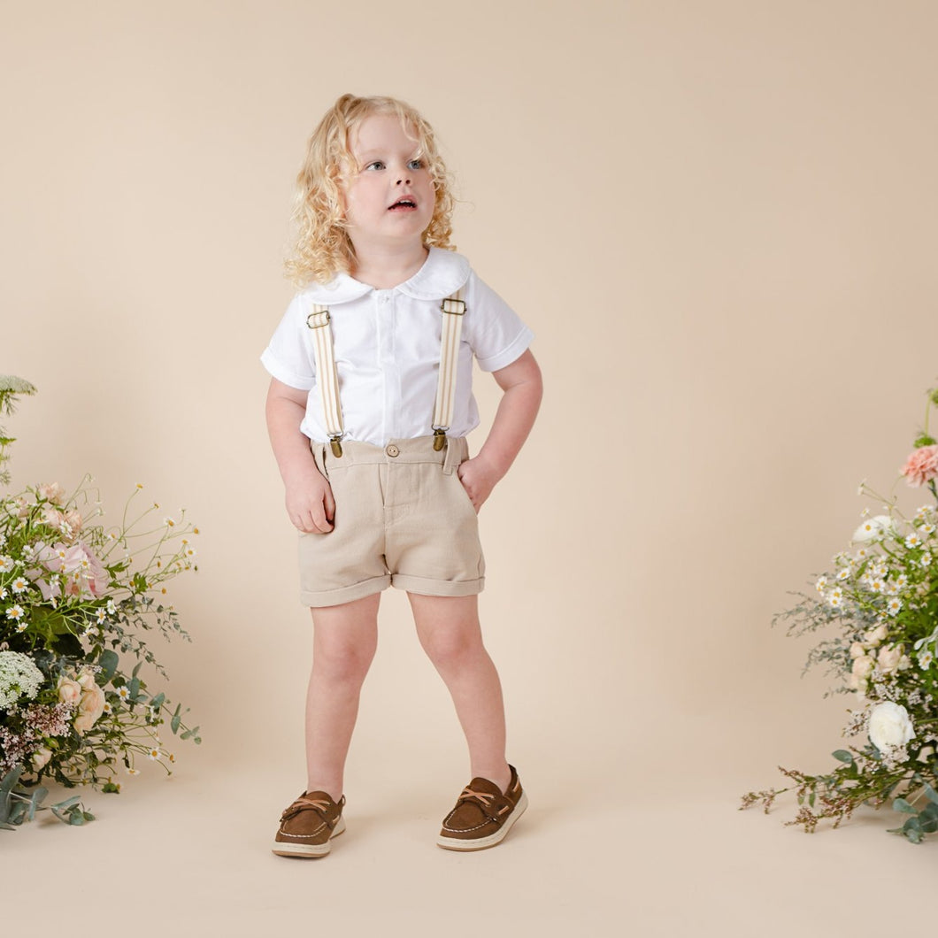 Beige cotton shorts with suspenders, faux fly and two pockets for baby boy or toddler boy on beige background.